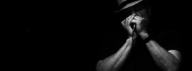 Black and white photo showing a man wearing a fedora and playing a harmonica into a microphone
