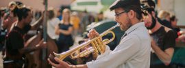 Man wearing white shirt, brown derby and glasses playing trumpet outside with orchestra while