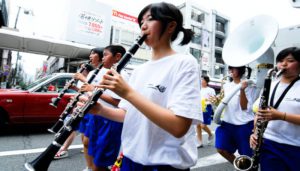 Rows of Asian kids playing various woodwind and brass instruments in a parade in the street