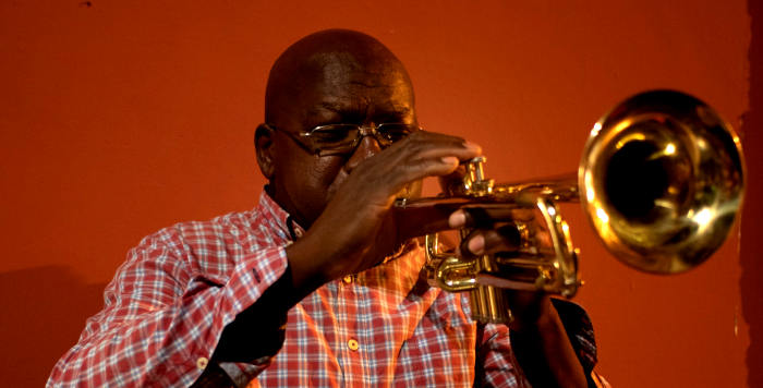 Black man wearing a red plaid shirt and playing trumpet on a red background